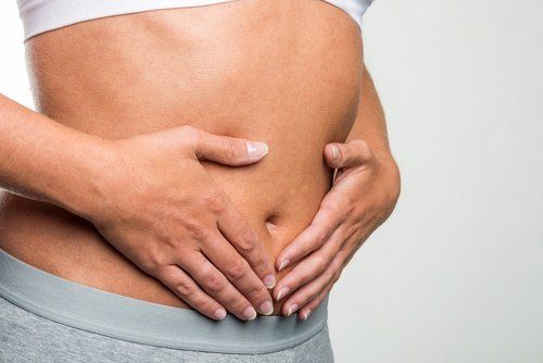 A person holds their lower abdomen with both hands, wearing a white top and grey pants against a plain background, possibly indicating their journey towards achieving a healthy weight.