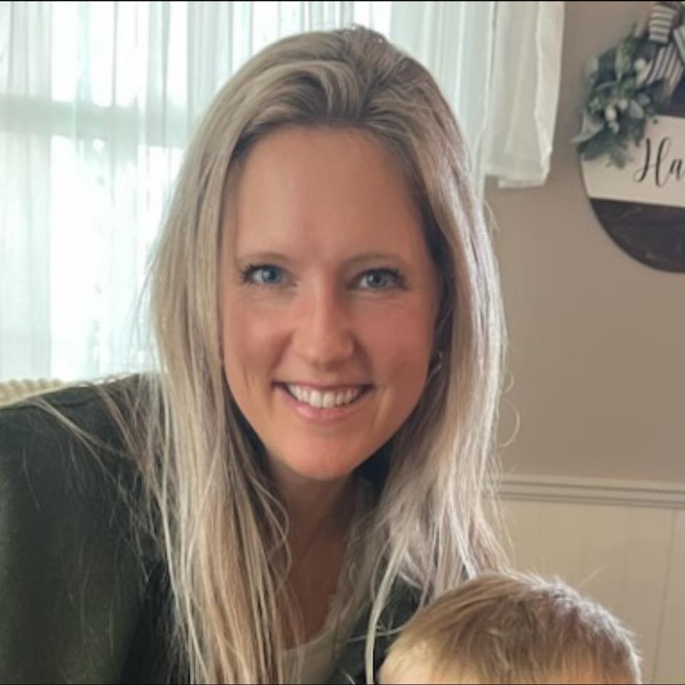 A woman with long blonde hair smiles at the camera while a child stands in front of her, slightly out of focus. The background includes a window with curtains and a wall adorned with home decor, evoking a sense of holistic harmony.