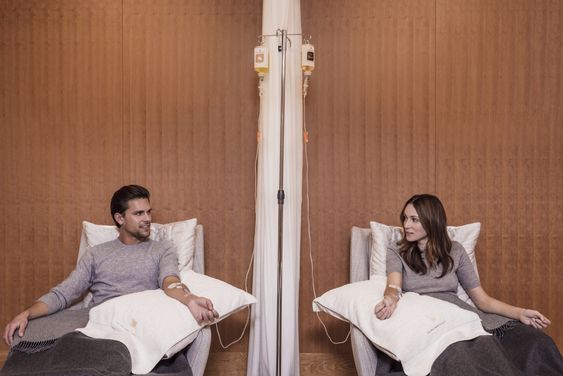 Two individuals sit in separate chairs with IV drips attached to their arms, facing each other in a room with a wooden wall. Both are dressed casually and appear to be engaged in conversation, perhaps discussing the benefits of hydration or energy through these vitamin-infused treatments.