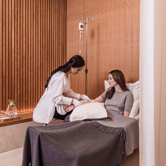 A woman is seated on a lounge chair, receiving an intravenous vitamin injection administered by a healthcare professional in a room with wooden walls. She is covered with a gray blanket.