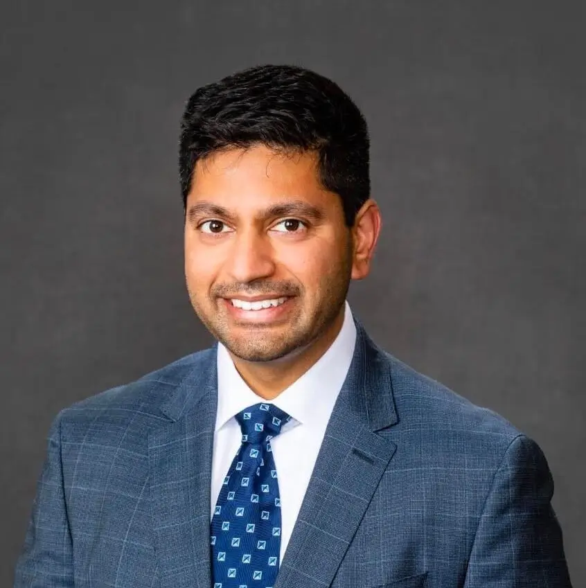 Man in a blue checkered suit with a patterned blue tie, smiling in front of a plain dark grey background, radiating optimal health.