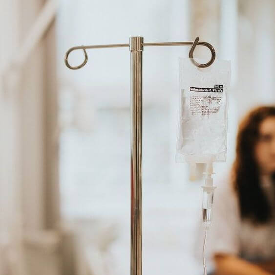 An IV drip bag hangs on a metal stand indoors, exemplifying a holistic approach, with an out-of-focus person seated in the background.