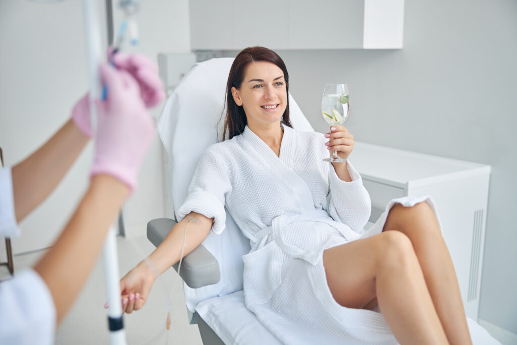 A woman in a white robe sits in a clinic chair receiving an IV treatment, holding a glass of water, while a medical professional adjusts the IV line.