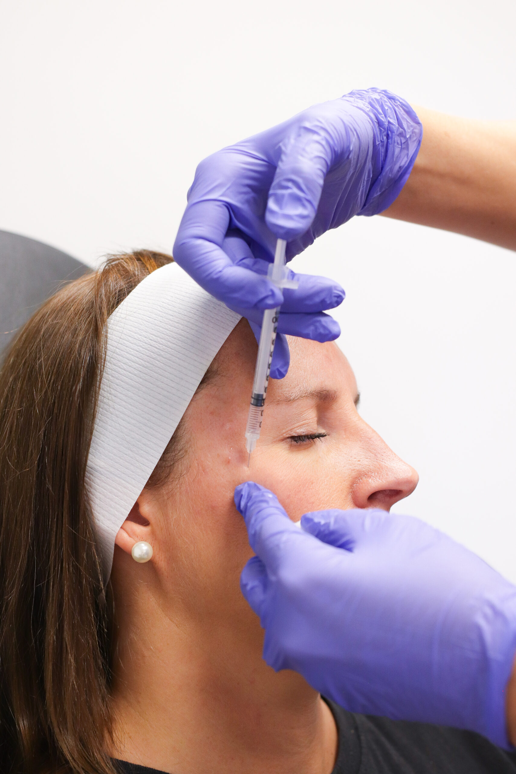 A person wearing a headband receives an injection on the side of their face from someone in purple gloves, part of a hormone therapy regimen focused on health and wellness.