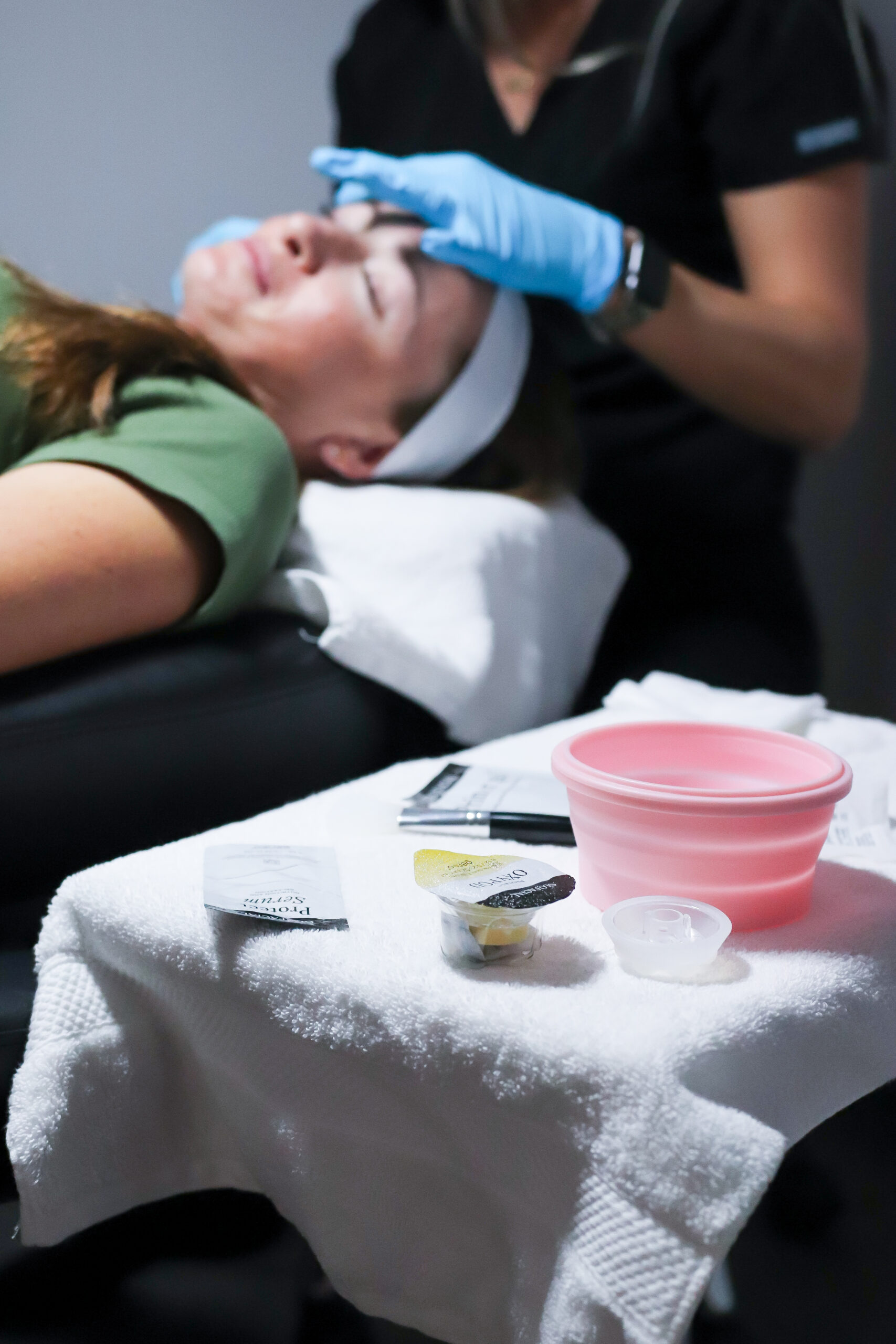 A person receives luxurious facial services while lying on a table, with a pink bucket and several skincare products arranged on a nearby towel-covered surface.