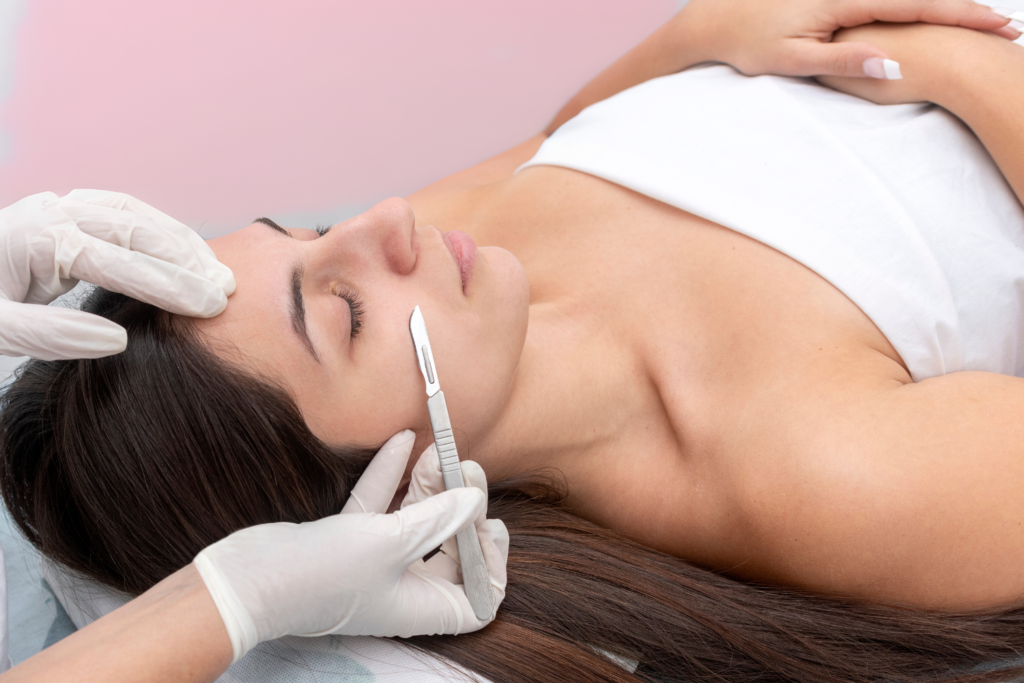 A woman lies on a treatment table with her eyes closed while someone in gloves holds a scalpel near her cheek.