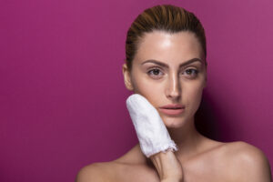 A woman against a pink background holds a white towel to her face, with her hair pulled back.