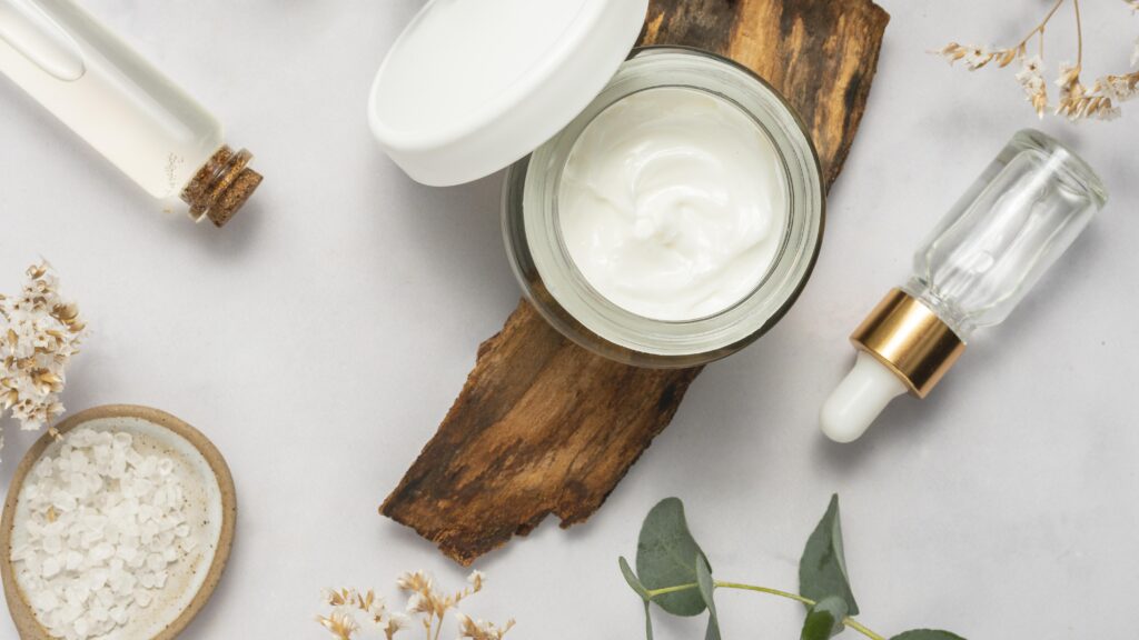 Open jar of white cream on wood slice with surrounding skincare items: small bottles, salt dish, dried flowers, and green leaves on a light surface.