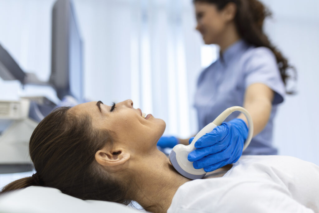 A woman is undergoing an ultrasound examination of the neck performed by a healthcare professional wearing blue gloves.