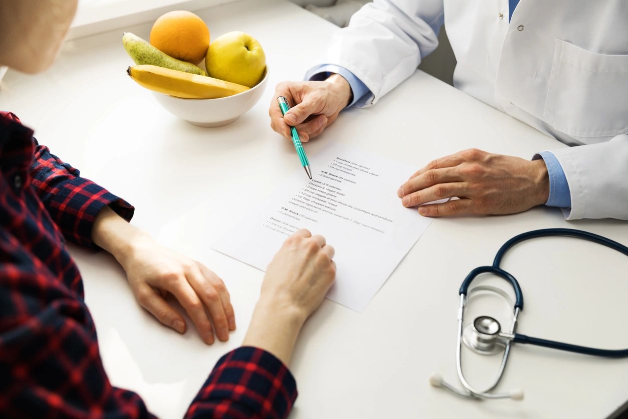 A doctor and patient discuss a medical document at a white table with a stethoscope and fruit bowl nearby.