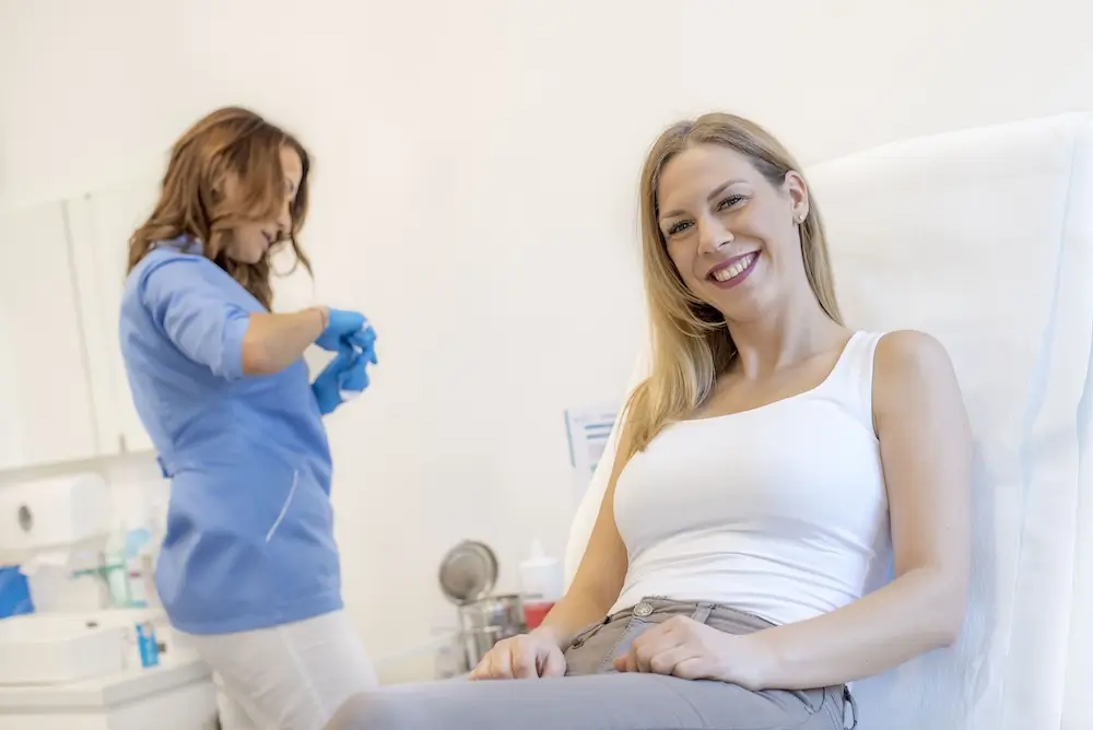 A woman sits and smiles in a medical exam room, anticipating her Hormone Replacement Therapy session, while a healthcare professional prepares equipment in the background.